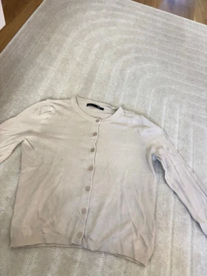 Beige cardigan från 157 - Ljusbeige cardigan med rund halsringning och knappar framtill från 157. Tröjan har långa ärmar och ribbade muddar vid ärmslut och nederkant. Perfekt att slänga över en t-shirt för en enkel och clean look. Storlek L men blivit lite mindre i tvätten så skulle säga S/M