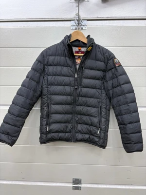 Svart dunjacka från Parajumpers - Snygg svart dunjacka i storlek youth medium från Parajumpers med quiltad design och hög krage. Jackan har två dragkedjeförsedda fickor fram och en patch med logga på ärmen. Perfekt för kalla dagar och riktigt lätt att bära. Klassisk och stilren look.