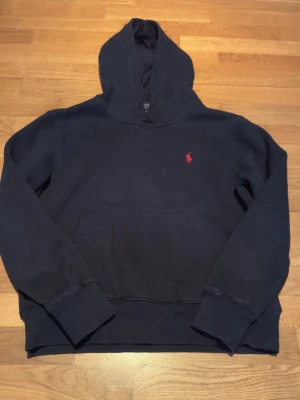 Polo Ralph Lauren hoodie - Mörkblå hoodie från Polo Ralph Lauren med klassisk röd logga broderad på bröstet. Hoodien har en stor känguruficka framtill, ribbade muddar vid ärmslut och nederkant samt en skön huva. Tillverkad i mjuk bomullsblandning som känns riktigt bekväm.