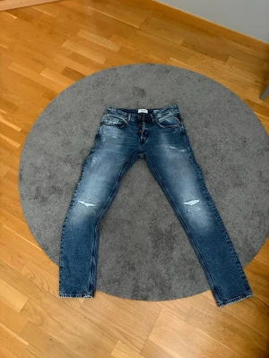 Replay jeans w29 - Säljer ett par blå jeans med slitningar och raka ben. Jeansen har klassisk femficksmodell, knappgylf och snygga slitdetaljer på framsidan. Materialet är denim i bomull och passformen är normal. Perfekta för en avslappnad och trendig look.W29 L32