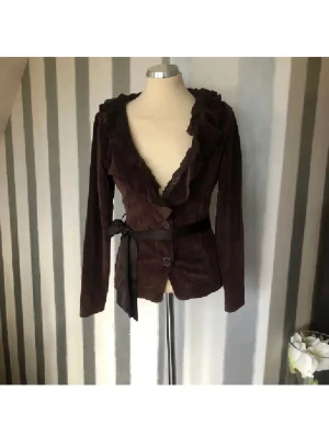 Brun sammet tröja med volangkant - Mörkbrun tröja i mjuk sammet med volangkant längs den djupa urringningen och knytband i midjan. Två knappar framtill och långa ärmar ger en elegant och trendig look. Perfekt för dig som vill sticka ut med en unik och snygg kavaj.