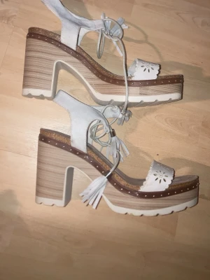 Chunky vita sandaler från Sixtyseven - Chunky sandaler från Sixtyseven i ljus mocka med höga klackar och platåsula i trälook. Sandalerna har vita snörningar med tofsar och blommönster på remmen över tårna. Perfekt för dig som gillar statement skor och cool design.