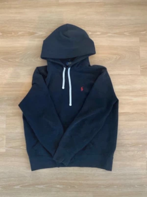 Svart hoodie från Polo Ralph Lauren - Säljer en svart hoodie från Polo Ralph Lauren med vit dragsko och klassisk röd logga broderad på bröstet. Hoodien har en stor huva och är tillverkad i mjukt material som är perfekt för chill dagar. Enkel och stilren design som funkar till det mesta.