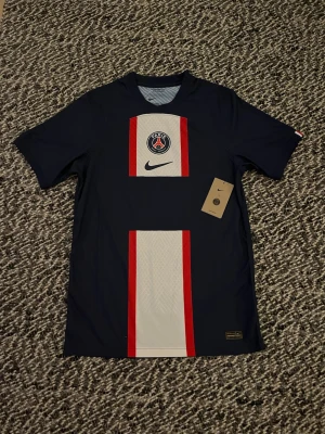 PSG matchtröja Nike blå/röd - Snygg PSG matchtröja från Nike i mörkblått med vita och röda detaljer på framsidan. Tröjan har klubbmärke och Nike-logga på bröstet samt korta ärmar. Tillverkad i lätt och ventilerande material, perfekt för fotboll eller träning.