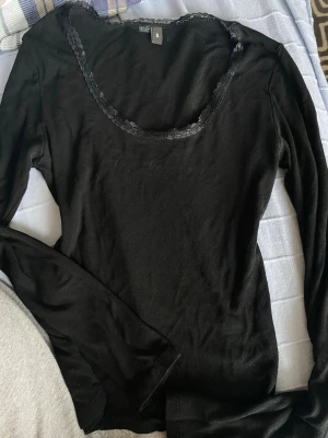 Svart långärmad topp med spetskant - Svart långärmad topp med rundad urringning och dekorativ spetskant runt halsen. Tunn och mjuk i materialet, perfekt för lager på lager eller som enkel bas. Enkel och stilren design som passar till det mesta.