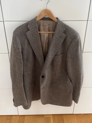 Grå tweedkavaj med klassisk passform - Stilren grå tweedkavaj med klassisk passform och två knappar framtill. Kavajen har breda slag, tre fickor och fyra mörka knappar vid ärmsluten. Materialet är grovt vävt och ger en tidlös look. Perfekt för dig som gillar en dressad men avslappnad stil.