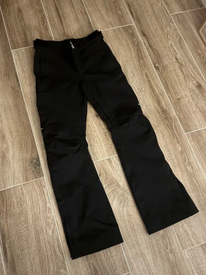 Svarta täckbyxor från Dope - Säljer ett par svarta täckbyxor från Dope med raka ben och dragkedja fram. Byxorna har diskreta fickor och en liten logga på benet. Perfekta för vinterns alla äventyr i backen eller snön.