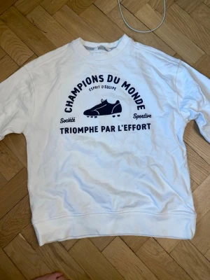 Vit sweatshirt från MNG med tryck - Vit sweatshirt från MNG med svart tryck på bröstet som har texten 'Champions du Monde' och en fotbollssko. Tröjan har rund hals, ribbade muddar och är gjord i mjukt material. Perfekt för dig som gillar sportiga och coola plagg.