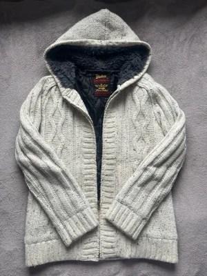 Vit/beige stickad cardigan - Mysig vit stickad jacka från Petroleum med kabelstickat mönster och dragkedja framtill. Jackan har en stor huva fodrad med mjuk grå fuskpäls och svart innerfoder. Perfekt för kyliga dagar och riktigt skön att bära.