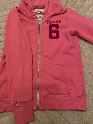 Rosa hoodie från Superdry Osaka - Rosa hoodie från Superdry med dragkedja, huva och fickor framtill. Tryck med texten 'OSAKA 6' i mörkrosa på bröstet. Mjuk insida och ribbade muddar vid ärmar och nederkant. Perfekt för en avslappnad och sportig stil.