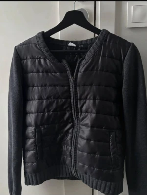 Svart quiltad cardiganjacka från Esprit - Snygg svart cardiganjacka med quiltad framsida och stickade ärmar. Jackan har dragkedja framtill, två fickor och ribbade muddar. Perfekt för lager på lager och ger en cool, modern look. Liknar den från moncler 