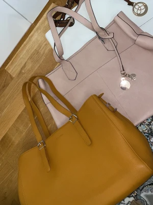 Två handväskor i skinnimitation - Säljer tre snygga handväskor. En är senapsgul med stilrena silverdetaljer och dubbla axelband från Don Donna, den andra är ljusrosa med dekorativ berlock och stora fack; och den tredje är ett stort vit färgade väska som jag tycker passar bäst på sommaren med sin stiliga berlock och dekorativa brun färg som kan hålla massa grejer i sig.  Alla tre är rymliga och har en klassisk, fyrkantig form i skinnimitation.