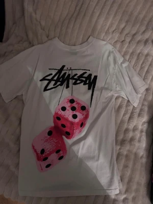 Stüssy Pink Dice T-Shirt - Vinted Dice Graphic - Säljer en snygg och populär Stüssy tröja i vitt med ikoniskt tryck av rosa tärningar och den klassiska loggan. Mjuk bomull, skön passform och perfekt för dig som gillar streetwear. Skick: Mycket bra, sparsamt använd Färg: Vit Detaljer: Rosa tärningsmotiv + svart Stüssy logo Storlek: S men är mer som M Passform: Lösare passform
