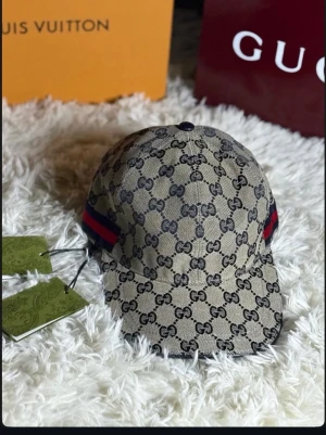Beige GG-mönstrad keps från Gucci - Säljer en beige keps från Gucci med det klassiska GG-monogrammet över hela kepsen. Kepsen har marinblått innerfoder och röda samt marinblå ränder på sidorna. Materialet är canvas och modellen har böjd skärm. Perfekt för dig som gillar exklusiva accessoarer.