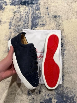 Mörkblå Christian Louboutin sneakers - Snygga mörkblå sneakers från Christian Louboutin med klassisk röd sula och coola nitar på tån. Skorna är i mocka och har vita gummisulor. Perfekta för dig som vill sticka ut med en lyxig och edgy stil. Kan gå ner vid pris vid snabb affär. Byten accepteras!