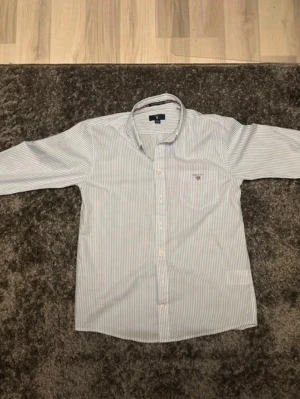 Randig skjorta från Gant i bomull - Klassisk vit skjorta med tunna blå ränder från Gant. Skjortan har lång ärm, knappar framtill och en liten broderad logga på bröstet. Tillverkad i mjuk bomull och har en normal passform. Perfekt för en clean och stilren look. 146/152 11-12.