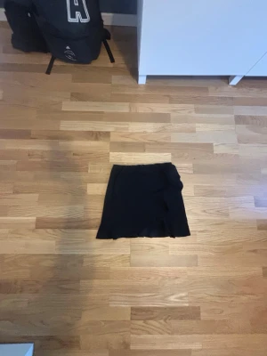 Svart kortkjol med volang - Säljer en svart kortkjol med snygg volangdetalj längs nederkanten. Kjolen har en omlott-look framtill och är perfekt för dig som gillar en enkel men trendig stil. Materialet känns mjukt och följsamt, vilket gör den bekväm att bära hela dagen.