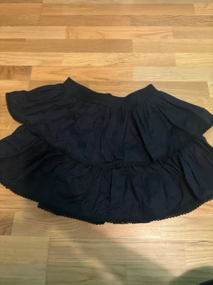 Svart volangkjol med spetsdetaljer - Säljer en svart kortkjol med dubbla volanger och fina spetsdetaljer längs kanterna. Kjolen har en luftig och flowy känsla, perfekt för dig som gillar en söt och enkel stil. Resår i midjan för skön passform.