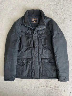 Svart parkajacka från Woolrich - Snygg parkajacka från Woolrich med hög krage och flera praktiska fickor framtill. Jackan har dragkedja och tryckknappar, samt en diskret logotyp på ärmen. Perfekt för kalla dagar och har en klassisk, tidlös look.