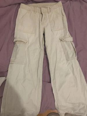 Beiga cargobyxor med fickor - Säljer ett par beiga cargobyxor med bred passform och flera stora fickor på sidorna och baktill. Byxorna har bälteshällor och stängs med dragkedja och knapp. Perfekta för en avslappnad och trendig streetwear-look.