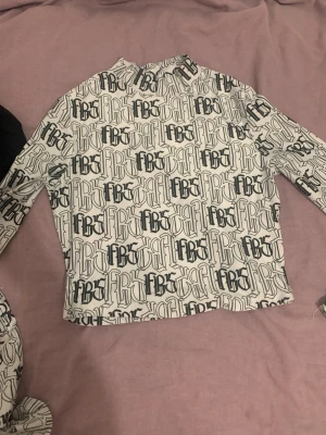 Vit långärmad topp med tryck - Trendig vit långärmad topp med svart grafiskt tryck över hela plagget. Hög hals och rak passform, perfekt att styla med jeans eller kjol. Materialet känns mjukt och stretchigt, vilket gör den bekväm att bära hela dagen.