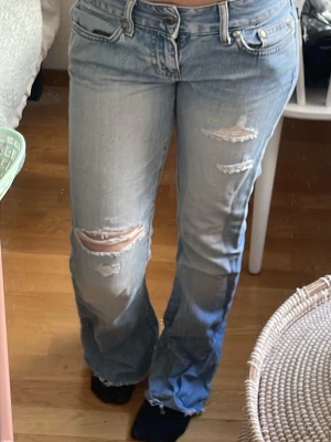 Ljusblå slitna bootcut jeans - Säljer ett par ljusblå bootcut jeans med slitningar och hål på låren och knäna. Jeansen har råa, fransiga benslut och en klassisk femficksmodell. Perfekta för en avslappnad och trendig look.