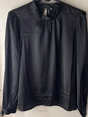 Svart satinblus med rund hals - Stilren svart blus  i glansigt satinmaterial. Blusen har rund halsringning, långa ärmar med lätt puff vid axlarna och knäppning bak i nacken. Perfekt för en chic och enkel look.