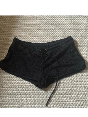 Svarta spets-shorts med snörning - Säljer ett par svarta lågmidjade shorts med snygg spetsdetalj och snörning i midjan. De har elastisk midja och kort passform, perfekta för varma dagar. Stilen är både chill och trendig, passar till många olika looks.