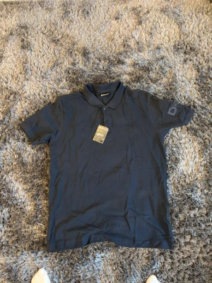 Mörkblå pikétröja från DKNY - Klassisk mörkblå pikétröja från DKNY med krage och knappar framtill. Tröjan har korta ärmar och diskret logga på ärmen. Perfekt till jeans eller shorts för en clean och stilren look.