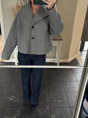 Grå oversized ullkappa - Stilren grå kappa i oversized modell med bred krage och två stora knappar framtill. Kortare längd och raka ärmar ger en modern look. Perfekt att slänga över en hoodie eller stickad tröja för en chill vibe. Materialet ser ut att vara ullblandning. Köpt på Vero Moda. 