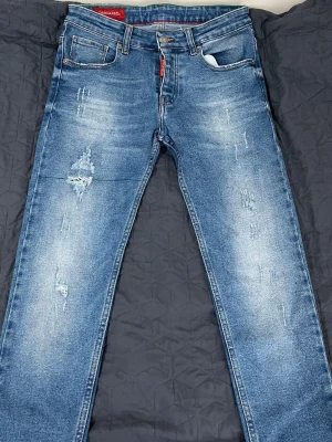 Blå slitna jeans från Dsquared2 - Säljer ett par blå jeans från Dsquared2 med snygga slitningar och distressed detaljer framtill. Jeansen har klassisk femficksmodell, raka ben och tydlig logga bak i midjan. Materialet är denim med tvättad look och coola slitningar.