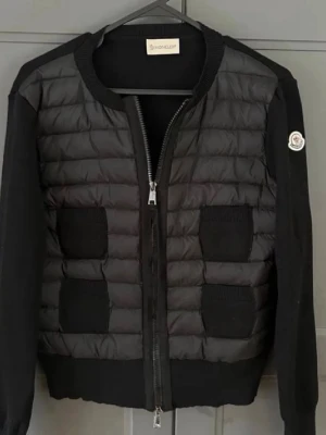 Svart cardiganjacka från Moncler - Snygg svart cardiganjacka från Moncler med quiltad front och stickade ärmar. Jackan har dragkedja, ribbade muddar och två framfickor. Moncler-logga på ärmen ger en exklusiv touch. Perfekt för lager-på-lager och streetstyle.