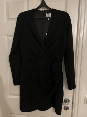 Svart omlottklänning med lång ärm - Stilren svart kortklänning med omlottdesign och lång ärm. Klänningen har en markerad midja med draperad detalj på sidan och v-ringad halsringning. Perfekt för dig som gillar en klassisk och elegant look.nypris 1600:- aldrig använd 