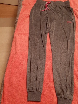 Gråa  yoga mjukisbyxor från Everlast - Mysiga grå mjukisbyxor från Lonsdale med rosa snörning i midjan och ribbade muddar vid bensluten. Tillverkade i mjukt material, perfekta för chill eller loungewear. Diskret Lonsdale-logga i rosa på ena benet.