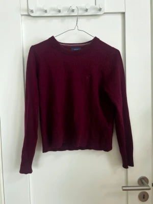 Vinröd stickad tröja från GANT - Mysig vinröd stickad tröja från GANT med rund halsringning och långa ärmar. Tröjan har en enkel, klassisk design utan mönster och är perfekt för lager på lager. Materialet ser mjukt och bekvämt ut, och färgen är djup och höstig. Den är väldigt liten k storlek skulle säga som xs