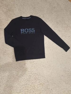 Svart långärmad tröja Hugo Boss - Snygg svart långärmad tröja från Hugo Boss med stort blått BOSS-tryck och HUGO BOSS-text på bröstet. Klassisk rund halsringning och stilren design som passar perfekt till jeans eller chinos.