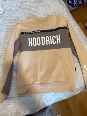 Beige sweatshirt från Hoodrich - Snygg beige sweatshirt från Hoodrich med kontrasterande brunt parti över bröstet och vit text. Rund halsringning och tryck på ena ärmen. Perfekt för en avslappnad streetwear-look.