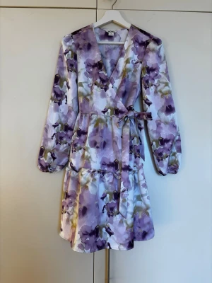 Lila/ vit ljuvlig blommig omlottklänning med volang från Monki storlek 32/ XXS - Ljuvlig kort omlottklänning från Monki med blommigt mönster i lila och gröna toner. Klänningen har långa ärmar, volangdetaljer och knytband i midjan. Perfekt skick! Storlek 32/ XXS