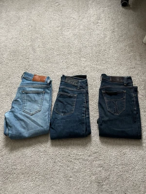 Tre par jeans i olika blå - Tiger of Sweden - Säljer tre par klassiska jeans i olika blå tvättar. Ett par är ljusblå med brunt lädermärke, de andra två är mörkare blå med svarta och grå läderpatches. Alla har raka ben och femficksmodell. Perfekta för en avslappnad och stilren look. Ljusblåa - 28/32. Mörkblåa med ljus stygn - 31/32. Mörkblåa med mörka stygn - 28/32.