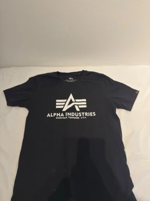 Svart Alpha Industries t-shirt - Snygg svart t-shirt från Alpha Industries med stort vitt logotryck på bröstet. Klassisk rund hals och korta ärmar. Perfekt för dig som gillar streetwear och vill ha en enkel men stilren look.