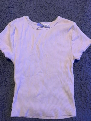 Ljusribbad t-shirt från H&M - En enkel och stilren ljus t-shirt från H&M med ribbad struktur. T-shirten har korta ärmar och rund halsringning, perfekt för en avslappnad look. Materialet känns mjukt och följsamt mot huden.