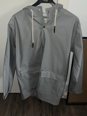 Grå regnjacka från 157 med huva - Snygg grå regnjacka från 157 med huva och vita dragsnören. Jackan har tryckknappar framtill och två stora fickor med lock. Perfekt för regniga dagar och enkel att matcha med olika outfits. Tillverkad i vattenavvisande material.