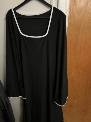 Svart långklänning med vit kant - En svart långklänning med vita kanter runt den fyrkantiga halsringningen och ärmsluten. Klänningen har långa ärmar och en enkel, rak passform. Perfekt för dig som gillar stilrena och minimalistiska plagg.