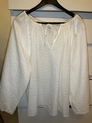 Vit strukturerad blus med knyt - Säljer en vit blus med strukturerat rutmönster och avslappnad passform. Blusen har lång ärm och en rundad halsringning med knytband framtill. Materialet känns luftigt och mjukt, perfekt för en trendig och bekväm look.