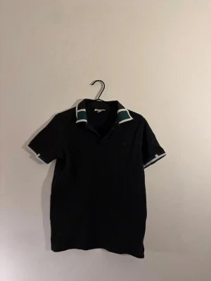 S Lacoste T-shirt - Klassisk svart pikétröja från Lacoste med gröna och vita detaljer på krage och ärmslut. Tröjan har korta ärmar och den ikoniska broderade krokodilen ton-i-ton på bröstet..