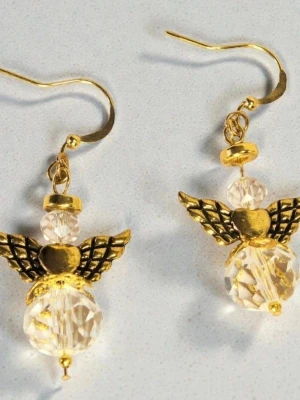 Guldiga ängelörhängen med pärlor - Unika örhängen i form av änglar med guldfärgade detaljer och vingar. Krokarna är guldpläterade. Örhängena har klara, facetterade glas pärlor som kropp och huvud, samt små hjärtan i mitten. Perfekta för dig som gillar statement-smycken med en söt och kreativ touch.