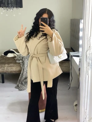 Beige kappa med knytskärp - Snygg beige Misay kappa med knytskärp i midjan och vida ärmar. Stilren och modern design, perfekt för att lyfta din outfit. Materialet ser ut att vara mjukt och bekvämt, och kappan har en avslappnad passform.