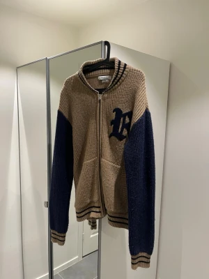 Beige stickad ziptröja med blå ärmar - Säljer en beige stickad ziptröja från Uran Outfitters med mörkblå ärmar och mörkblå broderad logga på bröstet. Tröjan har ribbade muddar och krage med svarta ränder samt två fickor framtill. Tröjan är knappt använd och är i storlek M