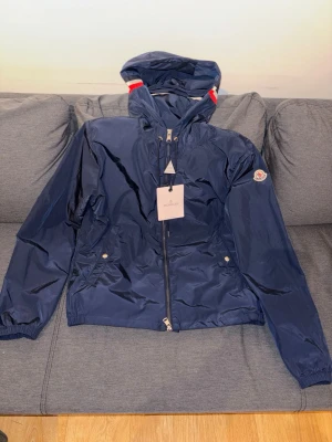 Marinblå vindjacka från Moncler - Snygg marinblå vindjacka från Moncler med huva och dragkedja framtill. Jackan har vita och röda detaljer på huvan, två fickor med tryckknappar och Moncler-logga på ärmen. Tillverkad i lätt, glansigt material som står emot vind. Perfekt för en sportig och trendig look.