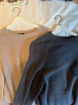 Beige och mörkgrå stickade tröjor - Två stickade tröjor, en i beige och en i mörkgrått, med rund hals och långa ärmar. Båda har enkel, klassisk design och är gjorda i mjukt stickat material. En för 250, två för 400!